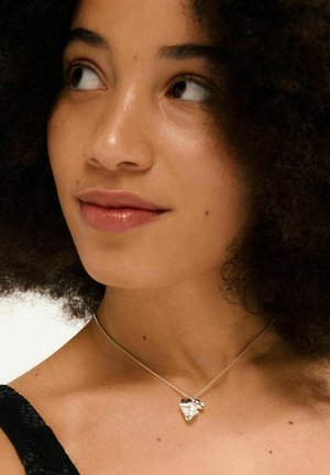Collana d'argento con pendente a forma di cuore, caratterizzata da una superficie lucida e un design intricato. Modella indossa un top in pizzo nero e ha i capelli ricci.