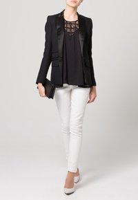 Schwarzer Blazer mit Satinrevers, darüber ein schwarzes Oberteil mit Spitzen-Details, kombiniert mit weißen Slim-Fit-Hosen und gemusterten High Heels.