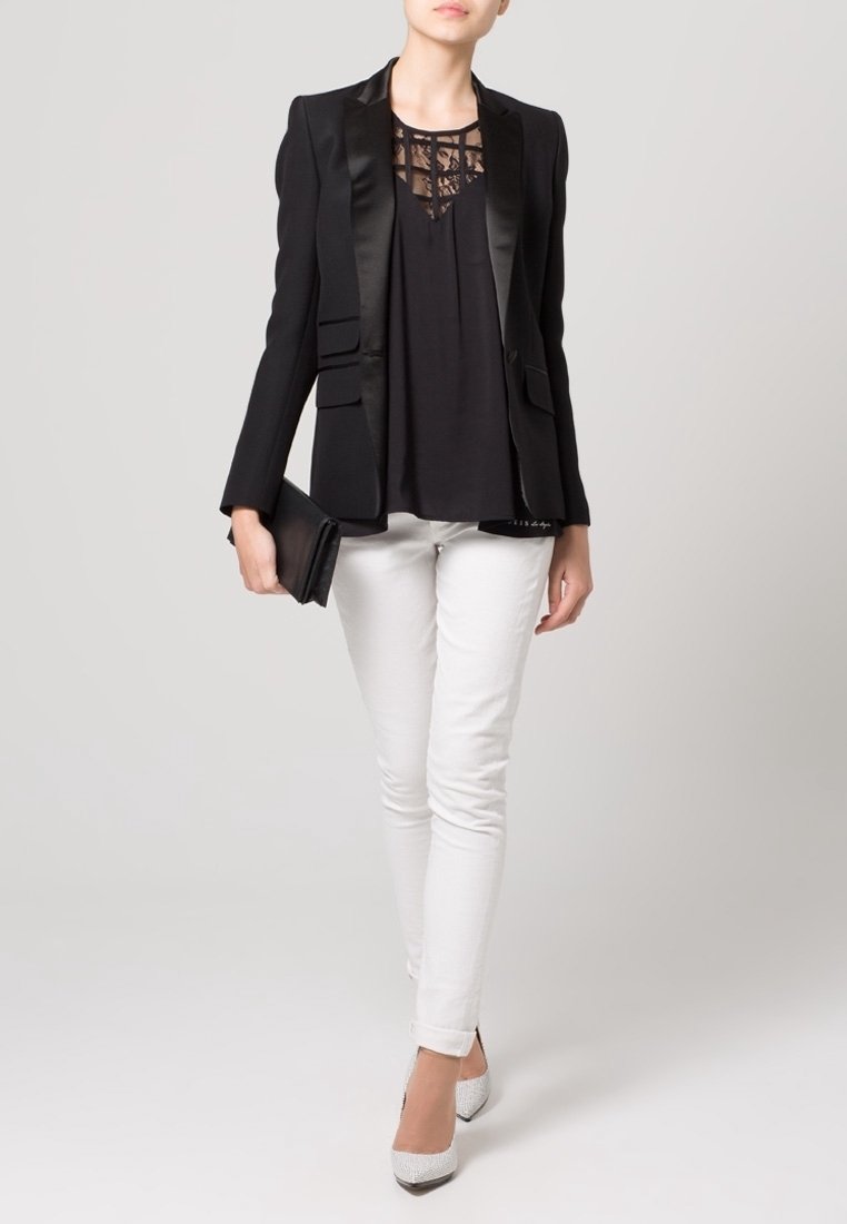 Schwarzer Blazer mit Satinrevers, darüber ein schwarzes Oberteil mit Spitzen-Details, kombiniert mit weißen Slim-Fit-Hosen und gemusterten High Heels.