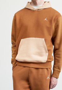Felpa con cappuccio marrone in pile, con tasca a marsupio beige, logo bianco sul petto e cappuccio di un colore più chiaro a contrasto; texture morbida, vestibilità rilassata.