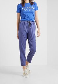 Blå t-shirt med en silverlogotyp, kombinerad med lätta, lila, tapered byxor med dragsko i midjan och svarta accenter. Vita sneakers.