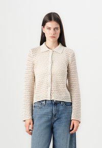 Beige gebreide cardigan met textuur, kraag, knoopsluiting aan de voorkant en lange mouwen, met een open patroon en een aansluitende zoom. Gemaakt met lichte blauwe jeans.