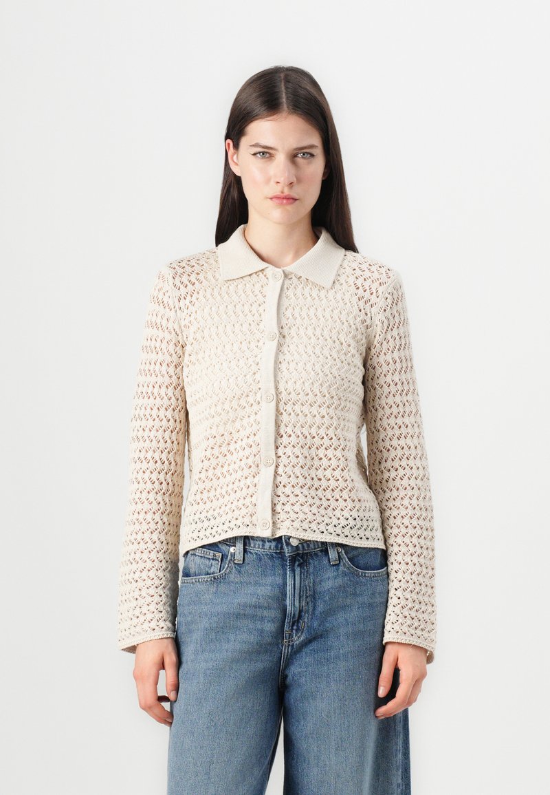 Beige gebreide cardigan met textuur, kraag, knoopsluiting aan de voorkant en lange mouwen, met een open patroon en een aansluitende zoom. Gemaakt met lichte blauwe jeans.