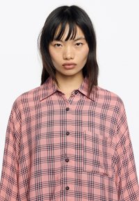 Camisa de cuadros rosas hecha de tela ligera, con un patrón de cuadrícula negra, cierre de botones, cuello y un bolsillo en el lado izquierdo.