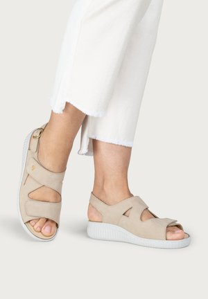 Feet wearing beige open-toe sandals with adjustable straps and white soles, paired with white cropped pants with frayed hems.