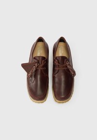 Um par de sapatos de camurça castanhos em estilo moccasin com solas de crepe, costura detalhada, cordões e etiquetas de marca num fundo simples.