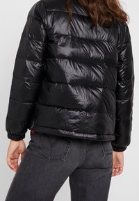 Giacca a vento nera con design trapuntato, finitura lucida e polsini elastici. Indossata con jeans scuri, mostrando una lunghezza cropped e un branding sottile.