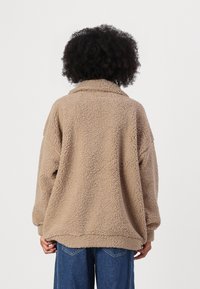 Béžový fleecový pulovr s límečkem, s textured povrchem a volným střihem. Ruka viditelná na spodní části, v kombinaci s modrými džíny.