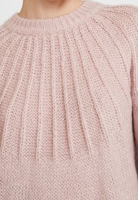 Jasnoróżowy sweter wykonany na drutach z prążkowanym wzorem i okrągłym dekoltem. Materiał jest miękki, z wyraźnymi pionowymi liniami i luźnym krojem.