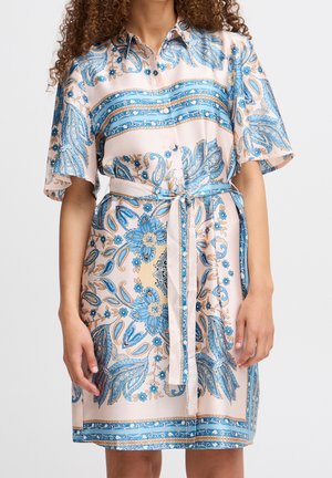 Femme portant une robe courte à manches courtes boutonnée avec des motifs floraux et cachemire bleus et beiges, nouée à la taille avec une ceinture en tissu assorti.