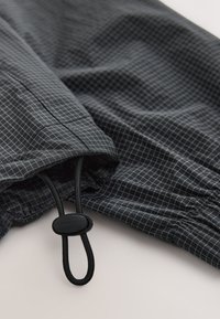 Čierna látka s jemným bielym mriežkovaným vzorom, s čiernou elastickou šnúrkou a plastovým zámkom na svetlom povrchu.