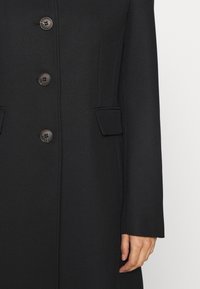 Black coat with a textured fabric, featuring five buttons, two front pockets, and a tailored design. Long sleeves with straight cuffs.