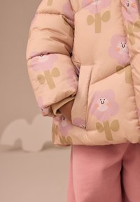 Pinke Steppjacke mit floralen Mustern in Hellviolett und Grün, mit einer Seitentasche. Weicher Stoff mit samtiger Textur, getragen über pinken Hosen.