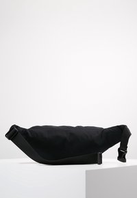 Musta olkalaukku pehmeällä, tyynytetyllä rakenteella; siinä on säädettävä hihna ja niittikiinnitys. Pehmeä pinta, minimalistinen muotoilu.