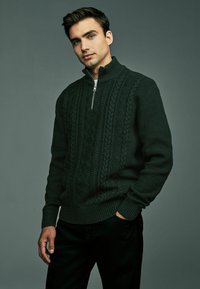 Suéter de punto verde oscuro con cuello alto, que presenta patrones de punto de cable y un cierre de media cremallera en la parte delantera. Combinado con pantalones negros.