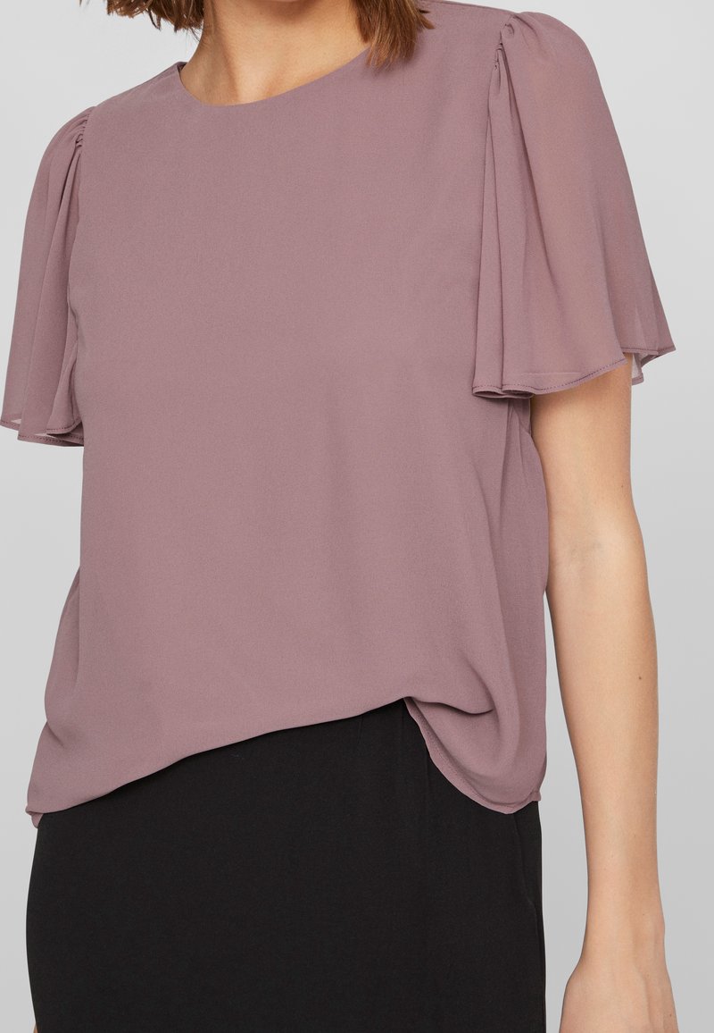Femme portant un chemisier mauve avec des manches courtes volantées et un col rond, rentré dans une jupe noire.