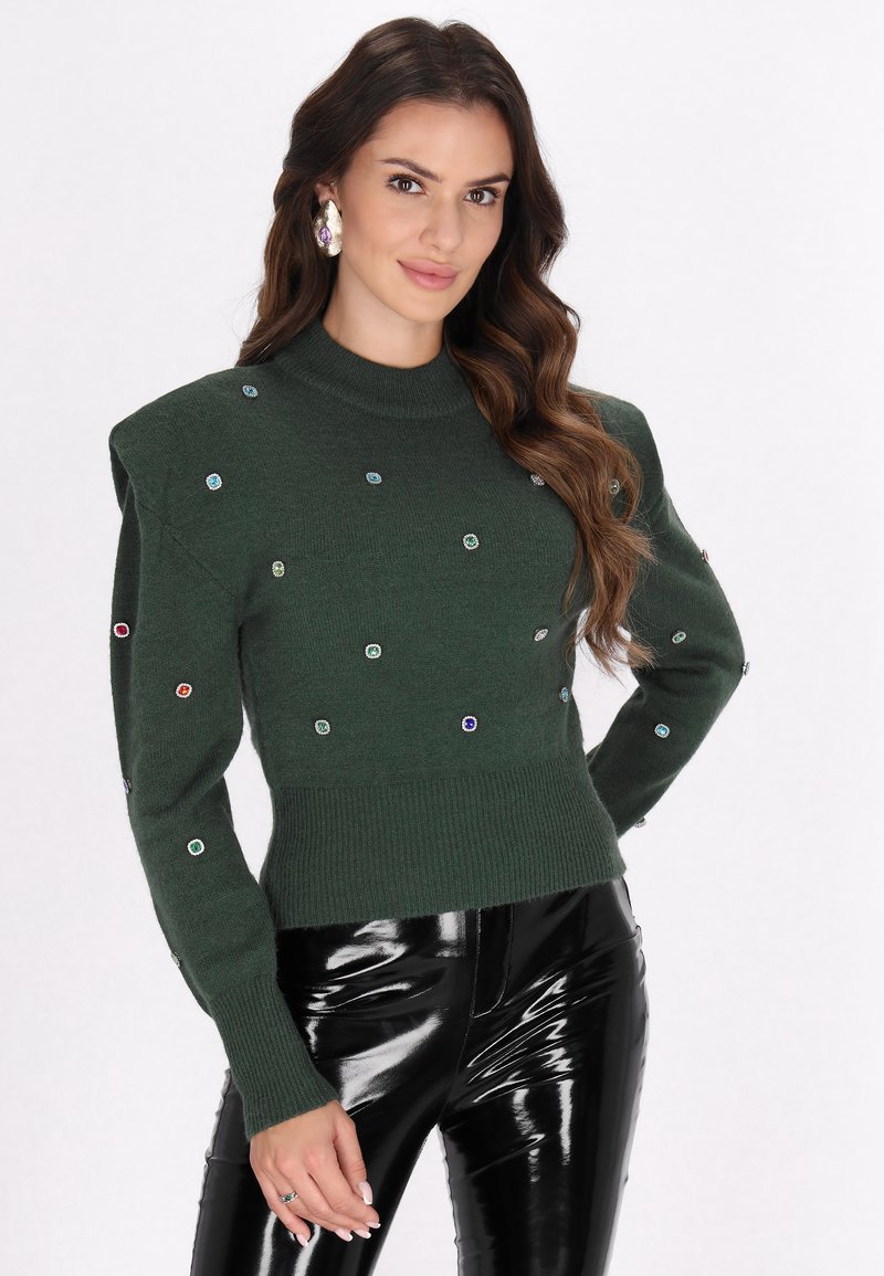 Groen gebreid truitje met opgeblazen schouderpartijen, voorzien van kleurrijke juweelversieringen en een geribbelde zoom, gecombineerd met glanzende zwarte vinyl broek.