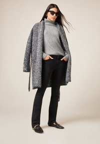 Suéter de cuello alto de punto gris, abrigo largo con estampado pata de gallo en blanco y negro, vaqueros negros con adornos y mocasines negros.