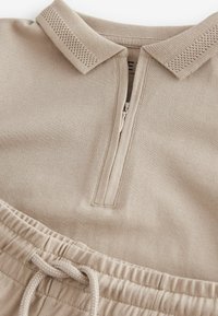 Beiges Polo-Oberteil mit einem Reißverschluss, strukturiertem Kragen und passenden Jogginghosen mit Kordelzug. Glatter Stoff mit einem dezenten gewebten Muster.