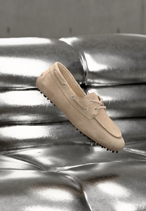 Beige suede loafer with lace detail and rubber studded sole placed on shiny metallic silver cushioned surface.