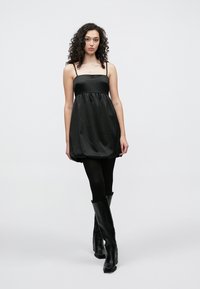 Μαύρο σατέν φόρεμα με λεπτές τιράντες, στενή μέση και φουσκωτή φούστα. Φορεμένο με μαύρες καλσόν και μπότες μέχρι το γόνατο για ένα ολοκληρωμένο σύνολο.