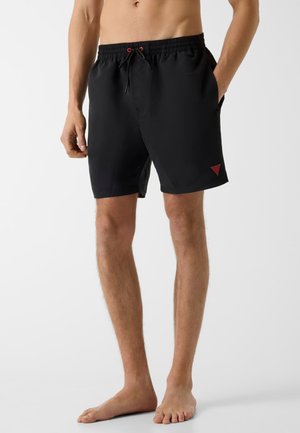 Man wearing black swim shorts with red logo and drawstring, standing barefoot against a plain light background.