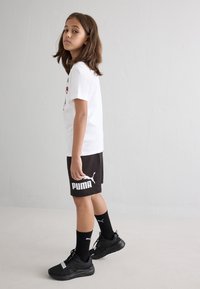 Παιδί που φοράει λευκό μπλουζάκι με γραφικά σχήματα, μαύρα σορτς με λευκό λογότυπο "PUMA", μαύρες κάλτσες και μαύρα αθλητικά παπούτσια.