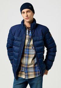 Chaqueta acolchada azul marino con cuello alto y costuras horizontales, llevada sobre una camisa de cuadros en beige, azul y marrón. Gorro de punto azul marino.
