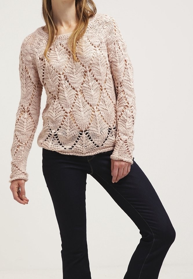 Femme portant un pull en tricot rose clair avec un motif en dentelle et un jean slim foncé, debout devant un fond uni.