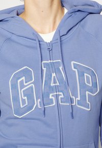 Hoodie zippé bleu clair avec un logo "GAP" blanc sur la poitrine. Comprend des cordons de serrage et un tissu texturé.