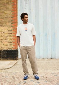 Kéz a zsebekben, fehér mintás pólót, bézs nadrágot és kék tornacipőt viselő férfi áll a macskaköves utcán, téglafalak és fa falak mellett.