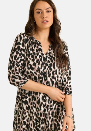 Kvinde med langt brunt hår iført en sort og beige leopardmønstret bluse med 3/4 lange ærmer og sorte knapper.