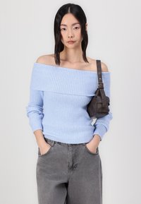 Lyseblå ribbet off-shoulder sweater parret med grå, baggy jeans. En lille, mørkebrun taske hænger over skulderen. Enkel, afslappet pasform.