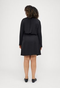 Abito nero con maniche lunghe, orlo fino al ginocchio, texture liscia. Indossato con scarpe nere basse, mostrato di spalle su uno sfondo neutro.