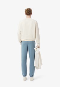 Huppari vaaleanbeige värissä, jossa ribbireunat hihoissa, yhdistetty sinisiin verkkareihin, joissa on joustavat nilkkaosat. Malli pitää kevyttä takkia.