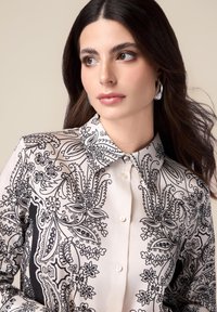 Blouse à manches longues en tissu crème avec un motif floral et paisley noir, dotée d'un col pointu et d'une fermeture à boutons.