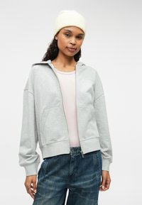 Ανοιχτό γκρι φούτερ με φερμουάρ και μαλακή υφή, μεγάλες μπροστινές τσέπες και εφαρμοστό τελείωμα, συνδυασμένο με ανοιχτό ροζ μπλούζα και μπλε τζιν παντελόνι.