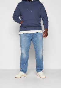 Felpa navy con tasca frontale, indossata sopra una maglietta bianca, abbinata a jeans azzurri chiari e sneakers crema con suole marroni.