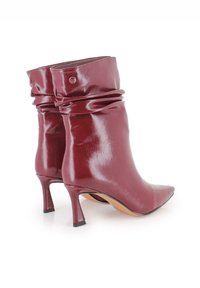 Botas de tobillo de charol rojo con diseño arrugado, punta afilada y tacón delgado de 6.5 cm. Presenta un detalle de logo grabado en la parte trasera.