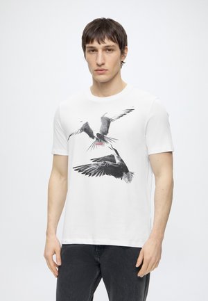 Νέος άνδρας που φοράει λευκό t-shirt με ασπρόμαυρα πουλιά και κόκκινο κείμενο «HUGO», σε συνδυασμό με μαύρο τζιν και απλό φόντο.