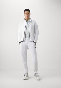 Hvid quiltet jakke med sorte lynlåse, parret med hvide sweatpants og grå-hvide sneakers. Lys blå polo shirt synlig. Glat tekstur.