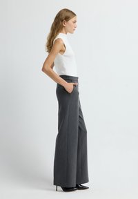 Top sin mangas blanco con textura acanalada; pantalones grises de pierna ancha y de talle alto; modelo mostrando perfil lateral, con las manos en los bolsillos, usando tacones negros.