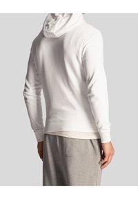 Felpa bianca con maniche lunghe, polsini a costine e cappuccio con cordino. Indossata sopra una camicia chiara, abbinata a pantaloni della tuta grigi. Texture liscia.