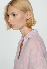 Camicia di lino rosa chiaro con una sottile texture e motivo a quadri, dotata di colletto e vestibilità rilassata, mostrata in un profilo laterale.