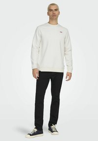 Witte sweatshirt met ronde hals en ribbels aan de mouwen, gecombineerd met zwarte jeans en marineblauwe sneakers met crèmekleurige veters. Minimale merknaam zichtbaar.