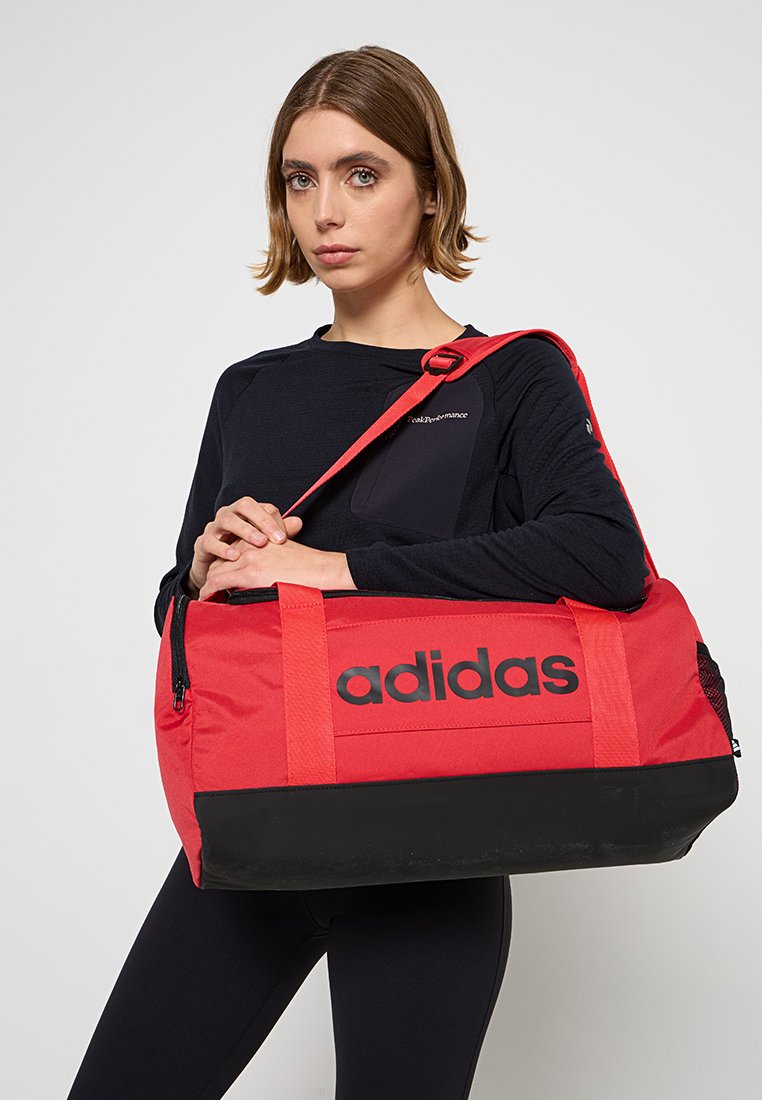 Bolsa de deporte roja con acentos negros, con un gran logo negro, asas resistentes y cierre de cremallera. El material parece duradero y con textura.