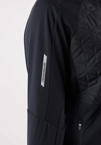 Giacca atletica nera con un pannello texturizzato sulla spalla con motivo geometrico. Include una tasca con cerniera e un logo riflettente sulla manica.