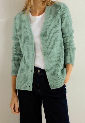 Femme portant un cardigan en grosse maille vert clair sur un t-shirt blanc et un jean foncé, main dans la poche, debout devant un mur clair.