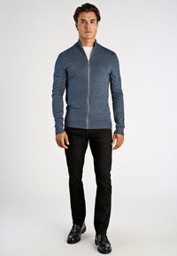 Man står klädd i en blå zip-up-tröja, vit undertröja, svarta byxor och svarta läderskor framför en enfärgad vit bakgrund.