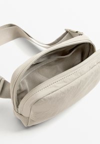 Bež kožni bum bag s zakrivljenim oblikom, zatvaračem na patentni zatvarač i mrežastim unutarnjim dijelom. Uključuje podesivu traku za sigurnu nošenje.